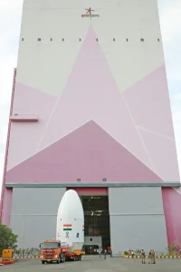 A white, ovular lander sits in the bed of an orange truck as it is taken out of a building painted with a pink triangle pattern.