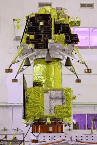 The Chandrayaan-3 lander, which has four legs sticking out to the side and looks like it is wrapped in gold foil, sits in a white-walled building.